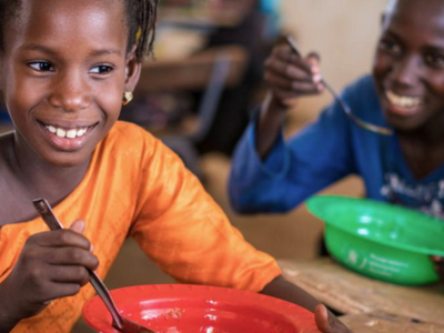 Child eating a meal from a red bowl in a classroom feeding programme, while another child eats in the background, highlighting school meals support.