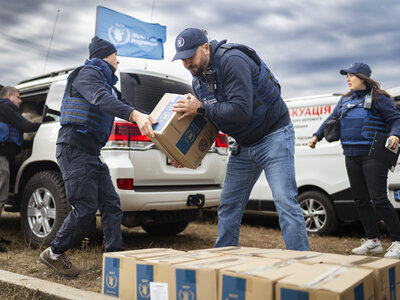 **Alt text:**   World Food Programme staff wearing protective vests unload and pass sealed boxes of assistance between vehicles in an open area, with multiple WFP-marked cars and a blue WFP flag visible in the background under a cloudy sky.