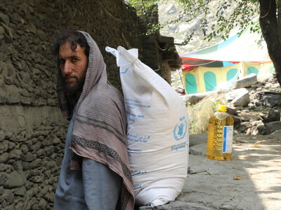 Photo: WFP/Arete/Muktar Nikrawa A person stands beside a large WFP food sack on a rocky path, with a bottle of cooking oil nearby and colourful tents visible in the background.