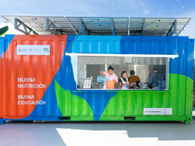 Three people inside a brightly coloured container with solar panels and signs promoting nutrition and education.
