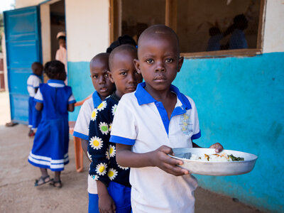 Children in school uniforms lined up outside blue-walled building; one child holds a plate of rice and vegetables, others wait in background.