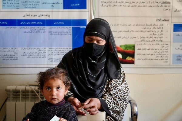 A caregiver sits beside a young child holding a nutrition packet in a clinic setting, with health information posters displayed on the wall behind them.
