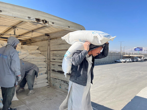 A person carries sacks of food from a truck during a roadside distribution, as others unload supplies under a clear sky in Afghanistan.