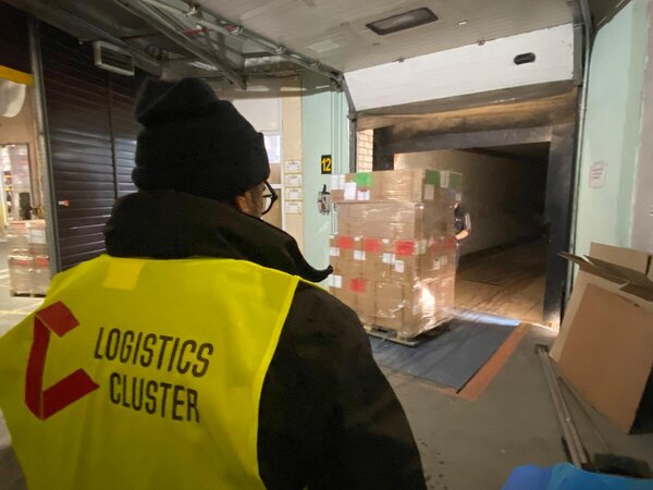 Logistics cluster worker in high-visibility vest oversees shrink-wrapped aid cargo being moved through a warehouse loading bay into a truck.