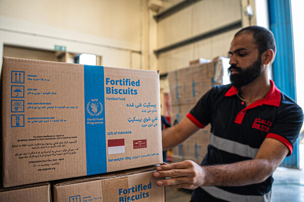 Worker stacks boxes of WFP fortified biscuits in a warehouse, with labelled cartons showing storage instructions and “Gift of Indonesia” text visible.