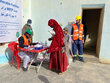 A woman in a head to toe red covering with a boy talks to a man sitting at a desk at an outtdoor WFP registration point