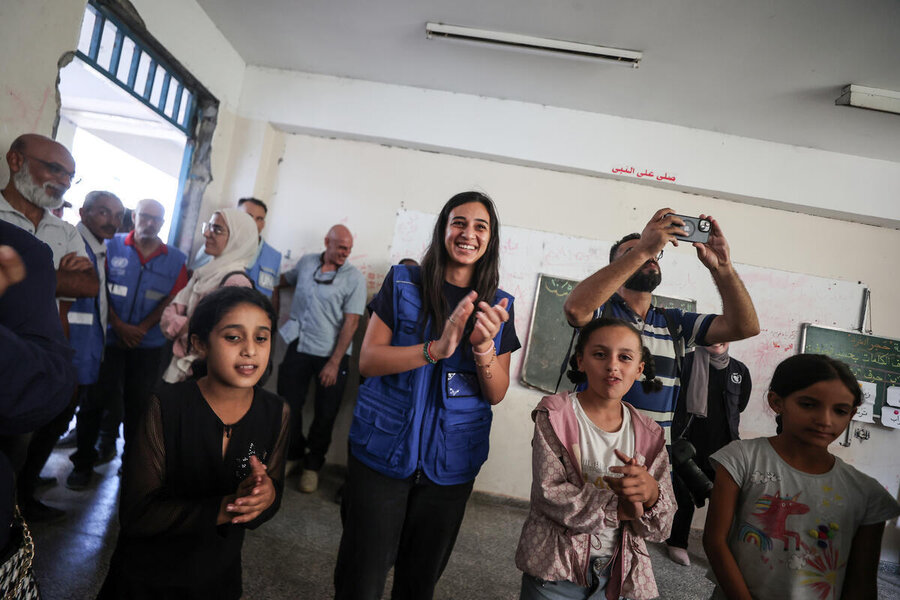 A young woman in a blue WFP vest stands in a classroom alongside cheering children