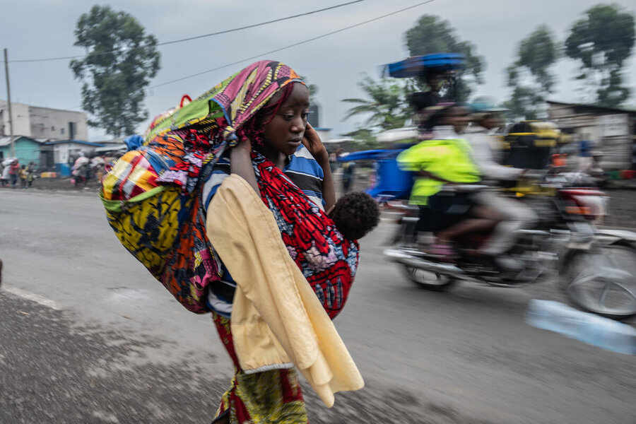 A woman with her belongings strapped to her back and a baby to her front walks down a road as a motorcycle passes