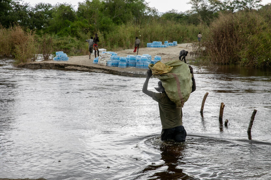 A man carrying a sack on this shoulders wades across a flooded road, packs of bottled water strewn on the otherside