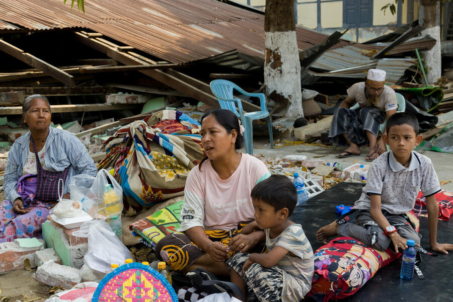 A women sat on the groun outdoors with a child, bags of food assistance, as another looks on in front of a damaged building