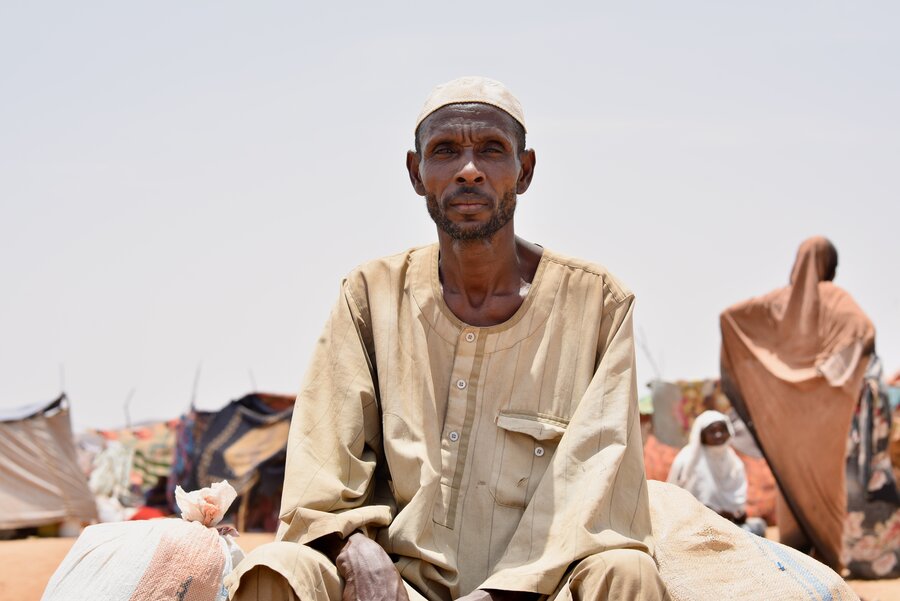 A man in a tan gown and white cap sits on bags, surrounded by sand, people and makeshift cloth tents. Photo: WFP/Mohamed Galal