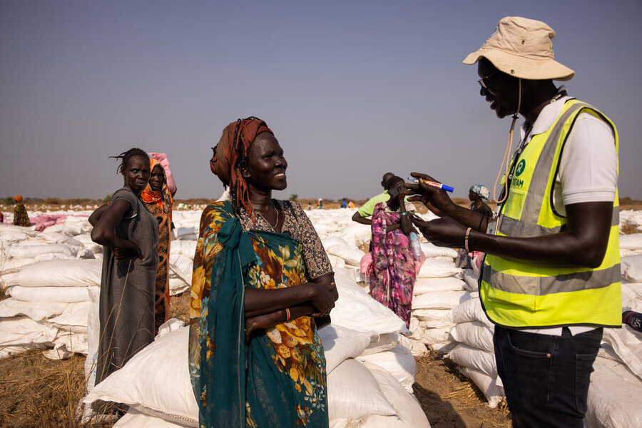 A WFP staff member in visibility vest speaks to a South Sudanese woman against a backhround of  white food bags