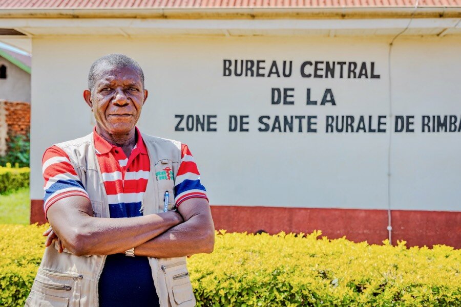 A man in a vest and colourful T-shirt stands in front of a health clinic in DRC's Ituri Province. Photo: WFP/Charly Kasareka