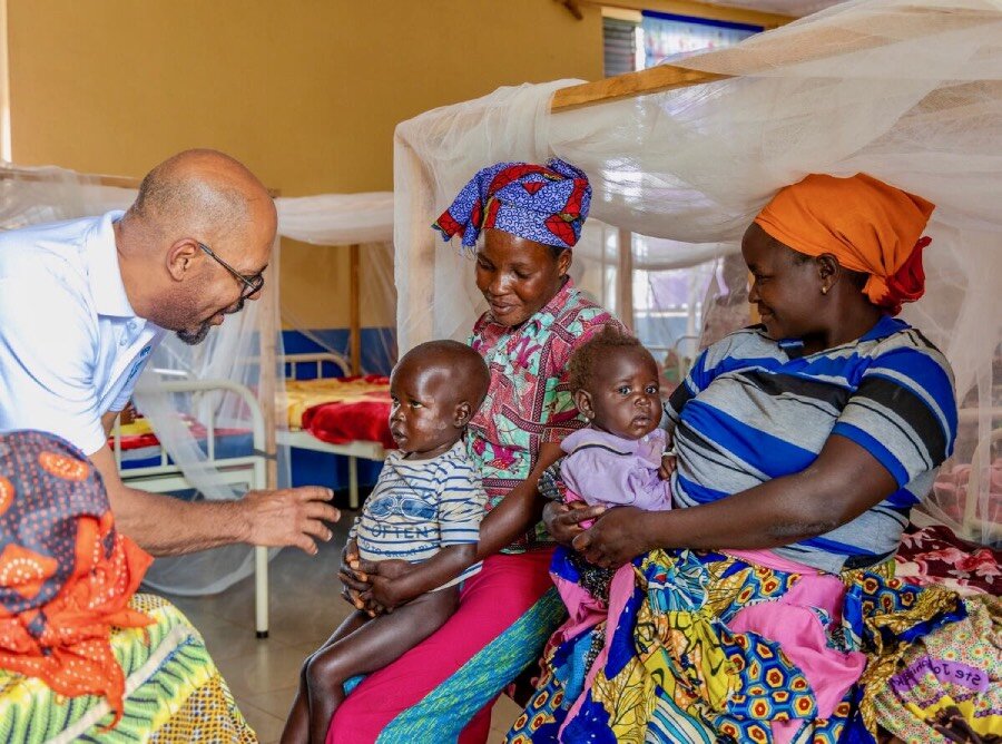A man in a white WFP T-shirt speaks to mothers and children at a health clinic. Photo: WFP/Charly Kasareka
