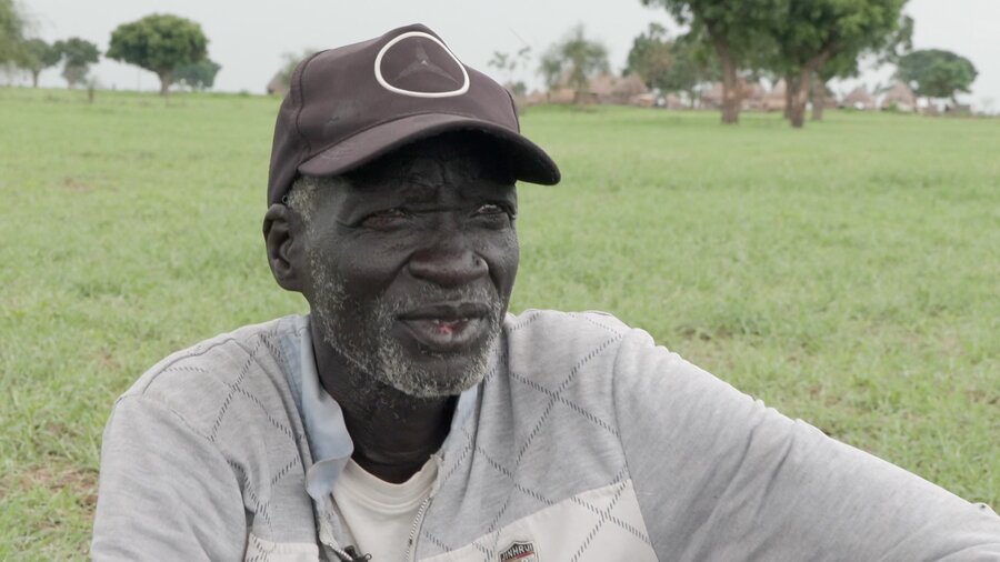 Closeup of an old man with a brown baseball cap and beige sweater sitting on a grassy plain with a few trees in the background. Photo: WFP/Peter Louis