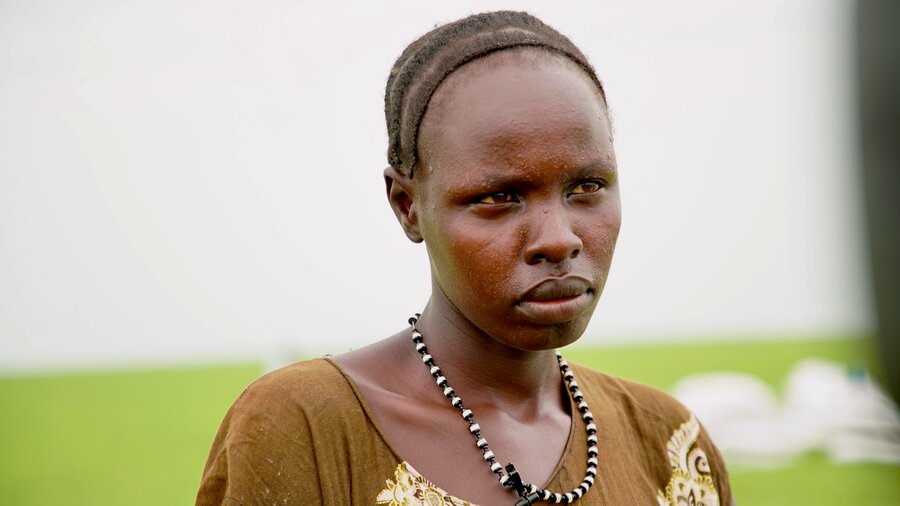 Closeup of a woman's face, which looks sad, backdropped by a green field and white food bags. Photo: WFP/Peter Louis