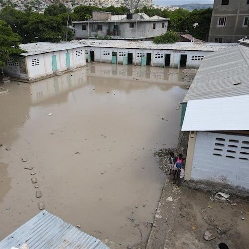Flooded courtyard surrounded by single-story buildings with two people standing near a wall.