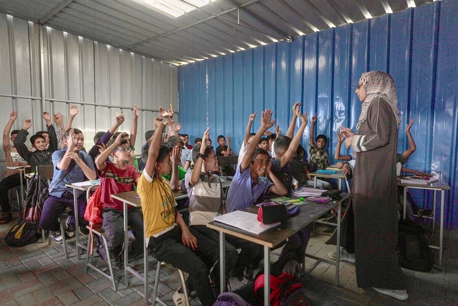 At a makeshift classroom in Gaza, a teacher in a grey gown and headscarf stands in front of a classroom of children with their hands raised. Photo: WFP/Jaber Badwan