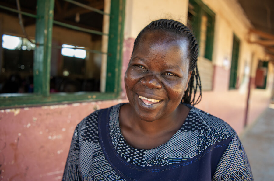 Closeup of a smiling woman in a dark patterned dress standing outside a low-level school building. Photo: WFP/Alessandro Abbonizio 