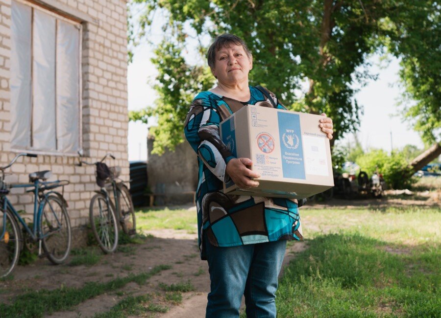A woman carrying a WFP food box stands in front of a brick house with bikes leaning on the side of it. Photo: WFP