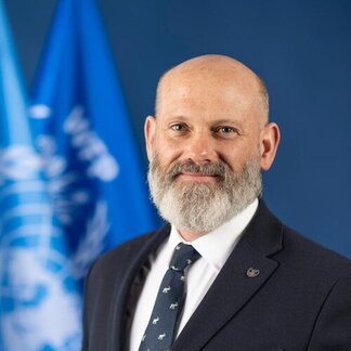 Man in a dark suit and patterned tie standing in front of blue United Nations flags on a solid blue background.