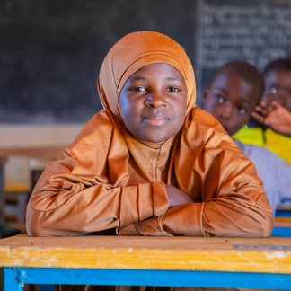 A student sits at a classroom desk, with a chalkboard and other students visible in the background.