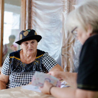 **Alt text:**   An older adult wearing a patterned hat sits at a table indoors while another person, partially out of focus in the foreground, counts or organizes banknotes. Plastic sheeting covers the wall behind them, suggesting a temporary or damaged setting, as cash assistance is being distributed.