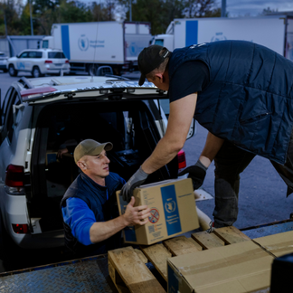**Alt text:**   Two individuals transfer sealed cardboard boxes marked with the World Food Programme logo from the back of a vehicle onto a platform, with additional WFP trucks and vehicles visible in the background, illustrating logistics support and aid distribution.