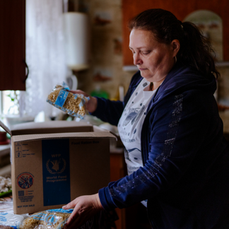 **Alt text:**   A person in a home kitchen removes packaged dry food items from a cardboard box labelled with the World Food Programme logo and places them on a counter, showing food assistance being received and organized indoors.
