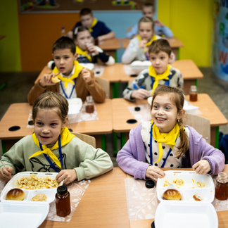 **Alt text:**   Children sit at desks in a brightly colored classroom, eating hot meals served in compartment trays with bread and drinks. More children are seated at desks in the background, illustrating a school meals setting.