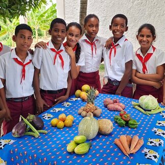 Students in uniform stand behind a table covered with colourful fruits and vegetables, including pineapple, carrots, and eggplants.