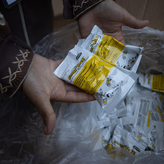 Close-up of two hands holding several small, rectangular packets of nutritional supplement. The packets are white with yellow labels featuring printed instructions and illustrations. More of the same packets are visible inside a large clear plastic bag in the background. Photo: WFP/Ali_Jadallah