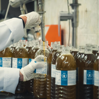 Close-up of a gloved worker filling bottles with oil using a dispensing nozzle. Multiple identical bottles with blue-and-white labels, bearing the World Food Programme logo, are lined up on a production surface in a food processing setting. Photo: WFP/Mostafa Ghroz