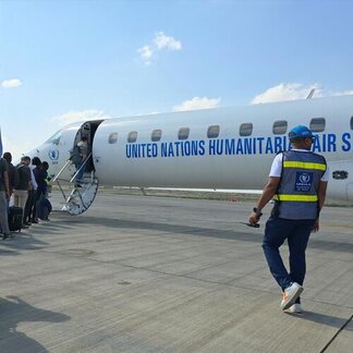 People board a UN Humanitarian Air Service plane on a tarmac, with a UNHAS staff member in a vest walking toward the aircraft under a partly cloudy sky.