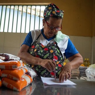 Person in a patterned apron writes on paper at a table with bags of beans and rice, in a kitchen with cooking oil bottles in the background.